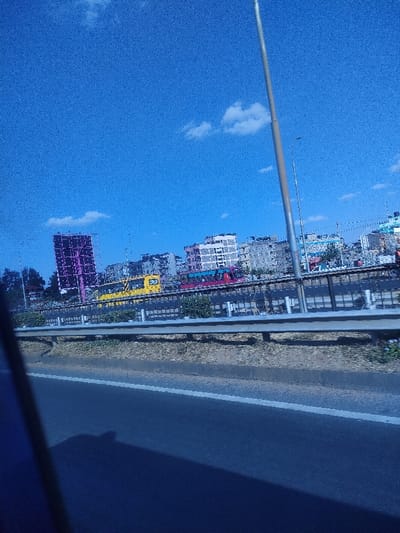 A roadside view under a clear, blue sky with scattered clouds. A safety barrier separates the road from an elevated area with greenery. Behind it, a cityscape emerges, featuring buildings of various heights and colors, including one with a distinctive purple hue. A yellow and a red bus are visible among the buildings. A tall, silver pole stretches into the sky on the right. The foreground is dominated by the road, partially obscured by shadows, suggesting the view is from within a moving vehicle.