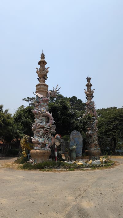 The image shows two tall, intricately decorated columns adorned with dragon sculptures. The columns are located on the side of a road in Lâm Sơn, Vietnam. The columns appear to mark an entrance or boundary to a property or area of interest. Other sculptural elements are visible near the base of the columns, along with a statue. The road is unpaved and runs alongside the columns. The sky is visible above the columns, suggesting an outdoor environment.