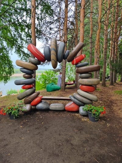 This media file depicts an outdoor scene in a wooded area near a body of water. The main subjects are large, artfully stacked, abstract stone sculptures arranged in a circular or frame-like formation. These sculptures are made of smooth, rounded, boulder-like objects in various earthy tones, including grey, brown, and red. Some of the stones have decorative stripes.
In the background, several people are visible. One person in a striped outfit stands near the sculptures. Further back, a person is relaxing in a hammock suspended between two trees. The setting is a forest with tall pine trees and lush green foliage. The ground is covered with dirt and fallen pine needles. In the distance, a lake or river can be seen, with a sandy shoreline.
Two potted plants with vibrant red flowers are placed at the base of the stone sculpture on the left and right sides, adding a touch of color to the scene. A wooden bench is positioned within the stone formation. The lighting suggests it is daytime, and the overcast sky hints at potentially cloudy or humid weather. The overall atmosphere appears tranquil and relaxed, with people enjoying the natural surroundings and the artistic installations. No text is visible in the media file.
