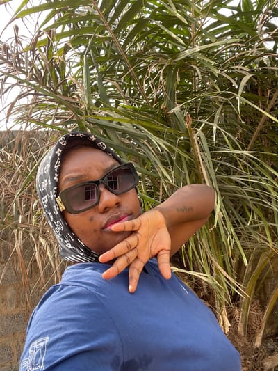 In Dzhidu, Nigeria, a headshot-style portrait depicts one individual with a dark skin tone, appearing to be an adult female, positioned centrally and looking directly at the camera. The individual wears a black headwrap patterned with white or light-colored monograms. Large black-framed sunglasses with dark lenses cover the eyes, featuring a gold-colored embellishment on the left temple. Pink lipstick is visible on the lips. A dark blue short-sleeved t-shirt, displaying a white graphic design on the left chest, is worn. The individual's right hand is raised, with fingers splayed, positioned near the chin. A small tattoo of text is visible on the inner left forearm. The background consists primarily of dense green foliage, with a light-colored block wall partially visible on the far left. The scene is illuminated by bright natural light, indicating daytime conditions.