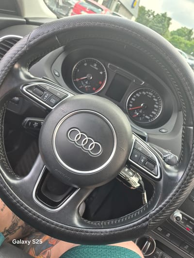 This media file shows a close-up view of the interior of a car, specifically the steering wheel and dashboard. The steering wheel is black and has the Audi logo in the center. There are several buttons on the steering wheel, including those for navigation and audio controls. The dashboard features a speedometer and a tachometer, both displaying readings. A car key is visible inserted into the ignition. The scene appears to be set during the daytime, with natural light illuminating the car's interior. The background shows glimpses of trees and buildings, suggesting an outdoor setting, possibly a parking lot or a street. The presence of tattoos on a person's leg, visible in the lower right corner, adds a personal element to the scene. The text "Galaxy S25" is overlaid on the image, likely indicating the device used for capture. The overall mood is neutral, focusing on the details of the car's interior and its immediate surroundings. While the exact location is identified as Chojnów, Poland, there are no specific visual cues in the image that confirm this definitively, beyond the general appearance of an urban or suburban environment. The image does not depict any specific activity or event, but rather a static view of the car's interior.