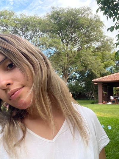 A person wearing a white shirt lies on grass with a large tree dominating the landscape in the background. Part of a building with a red-tiled roof is visible in the background behind the tree. The image is shot from a low angle, looking up at the tree and the person. The person's face is in focus, with their hair partially covering their face.