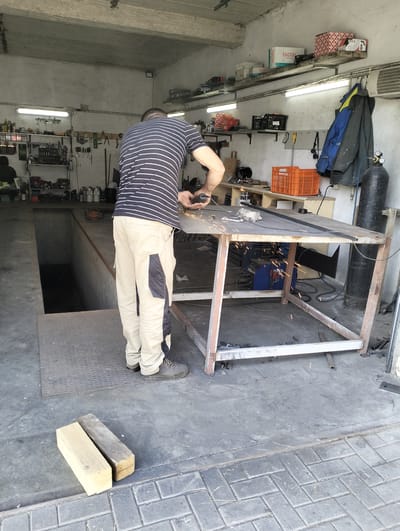 The image depicts a man in a striped t-shirt and beige work pants using a grinder on a metal table in a workshop located in Șoldănești, Moldova. Sparks are flying from the grinder, indicating an active metalworking process. The workshop environment includes shelves filled with tools and supplies, a welding machine, an orange plastic crate, and a large gas cylinder. A car pit is visible to the left of the man. The floor is concrete, with some interlocking pavers in the foreground. The lighting suggests it is daytime, with fluorescent lights also visible overhead. The overall scene is consistent with a functional, working garage or repair shop.