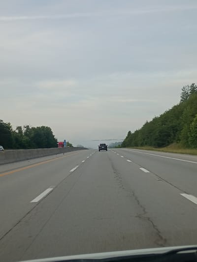 The image shows a view from inside a vehicle traveling on a highway in Mount Vernon, United States. The highway stretches ahead, with clear lane markings and some cracks in the asphalt. Trees line the left side of the road, creating a lush green border. In the distance, there is a hint of mist or fog, obscuring the landscape and creating a hazy effect.
A few vehicles are visible on the highway. A dark-colored SUV is driving ahead in the same lane. Further down the road, a red truck and possibly another vehicle are stopped or moving slowly near a sign. The sky is overcast, suggesting it might be early morning or late afternoon, or perhaps a generally cloudy day. The overall mood of the image is calm and serene, with the focus on the open road and the natural surroundings. No people are clearly identifiable, and the primary objects are vehicles and the road itself within a natural landscape.