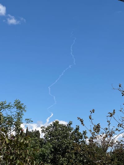 The image displays a daytime scene in Beit Yitshak Sha'ar Hefer, Israel, under a clear blue sky with scattered white cumulus clouds. A prominent, winding white smoke trail ascends diagonally from the lower-center of the frame, originating near or just above the tree line, and extends towards the upper-right, dissipating into the sky. This trail exhibits a distinct undulating or zig-zag pattern. The lower third of the photograph is occupied by the top canopy of several trees, featuring dense green foliage on the left and a mix of green and sparser, drier-looking branches on the right. No individuals or man-made structures are visible. The scene captures the visual aftermath of an object's trajectory through the atmosphere.