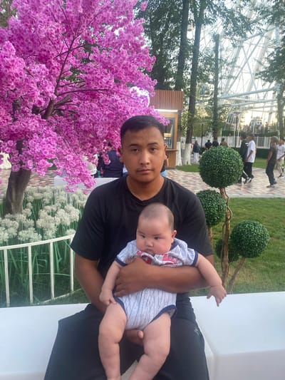 A man holds a baby in an outdoor park setting. The man, with short dark hair and a light complexion, wears a black t-shirt and black pants. He is seated on a white bench, holding the baby in his arms. The baby, wearing a striped onesie with red lettering, is facing the camera. In the background, a large tree with vibrant pink flowers is to the left of the frame. Behind the tree, there are white flowers and a fence. Further back, a Ferris wheel is visible, along with other people walking around the park. The ground is covered in grass and a paved walkway. The lighting suggests it is daytime, possibly late afternoon. The park appears to be well-maintained with various decorative plants and trees.