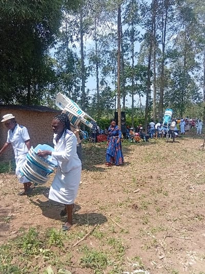 The scene depicts an outdoor gathering in Butali Chegulo ward, Kenya, under a clear sky during daylight hours. The ground is dry, reddish-brown earth with scattered green vegetation.

In the immediate foreground, two individuals, both predominantly dressed in white, are in motion. The person on the left wears a light-colored hat and a white short-sleeved top, carrying a stack of blue and white plastic basins. The person to their right, wearing a white long-sleeved dress, carries an identical stack of blue and white plastic basins. Both individuals are moving from right to left.

In the midground, a low-rise, light-brown building with a dark roof is visible on the left. To the right, a person in a blue patterned dress stands near a large black loudspeaker mounted on a stand. Further back, a crowd of people is gathered, many dressed in white attire. Multiple individuals within this crowd are observed carrying stacks of white plastic chairs, some balanced on their heads or shoulders.

The background is dominated by a dense stand of tall, slender trees, consistent with eucalyptus, reaching high into the bright blue sky with minimal cloud cover. A cluster of bamboo is visible on the far left. The collective activities suggest preparations for or participation in a community event.