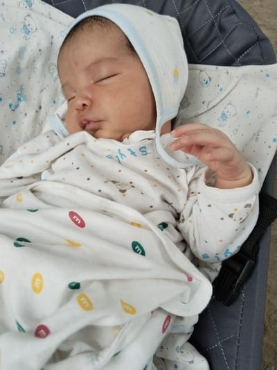 A newborn baby is sleeping soundly in a gray fabric bassinet, which is situated on a patterned fabric surface that resembles a mat or blanket. The baby is wearing a white onesie with a pattern of small stars and round shapes in various colors, including red, yellow, green, and blue, some of which appear to be stylized "M"s. The baby also wears a white bonnet with a light blue trim. The baby's right hand is visible, resting loosely. The background consists of the quilted gray fabric of the bassinet and the patterned surface underneath. The lighting suggests an indoor setting. The location in Nematjan, Uzbekistan, is not discernible from the image itself.
