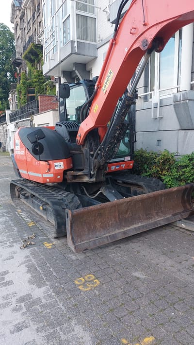 This is a close-up, eye-level, full-frame photograph of an orange and black compact excavator parked on a paved surface in Namur, Belgium. The excavator is the primary subject, positioned diagonally in the frame, with its blade facing the viewer. The background consists of a building with a grey facade and several windows, and on the left, a more ornate building with stone accents and ivy-covered balconies. The paved surface is made of interlocking grey pavers, some of which are marked with yellow spray-painted numbers, specifically "23" and "63." The excavator has a visible serial number "KX080-4" and logos for "Infra" and "Luyckx." The environment appears to be an urban street or construction site. No people are visible in the frame. The lighting suggests it is daytime, with overcast conditions.