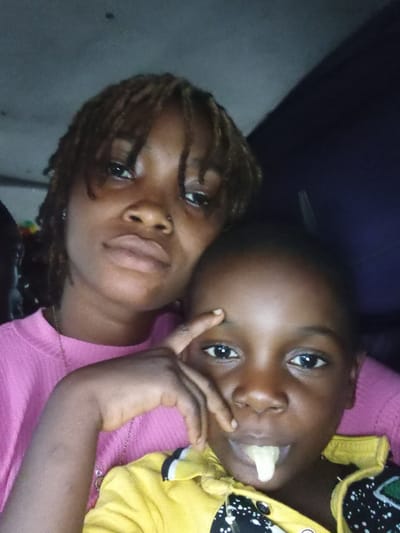 This image captures two individuals, an adult female and a child, in a close-up, diagonal portrait, likely taken indoors in Oba-Ile, Nigeria. Both subjects have dark skin and are looking towards the camera.
The adult, positioned slightly behind and above the child, wears a pink top and has dark hair styled in dreadlocks or braids. Her expression is neutral to thoughtful. A thin chain is visible around her neck. Her arm is gently placed around the child, with one hand resting near the child's eye.
The child, in the foreground, wears a yellow garment with black and white patterned sleeves. A small, light-colored piece of food, possibly a banana or plantain, is held in their mouth. The child has wide, dark eyes and a neutral expression.
The background is dark and indistinct, suggesting an intimate indoor setting with subdued lighting, possibly indicating evening or a room with limited natural light. The overall scene conveys a sense of warmth and closeness between the two individuals. There is no visible text in the image.