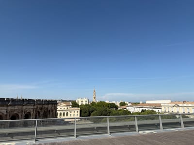 The image displays a panoramic view of the city of Nîmes, France, captured from an elevated vantage point. In the foreground, a modern wooden deck with a glass railing frames the scene. Beyond the railing, an ancient stone amphitheater, identified as the Arènes de Nîmes, dominates the left side of the image with its weathered arches and battlements. In the mid-ground, a cluster of buildings, including a tall church spire, are nestled amongst lush green trees, hinting at the city's urban landscape. The background features more buildings with terracotta roofs and architectural details consistent with French cityscapes. The sky is a clear, bright blue, suggesting a sunny day. No people are distinctly visible in the image, nor are there any observable interactions. The overall scene conveys a sense of historical depth juxtaposed with modern infrastructure.