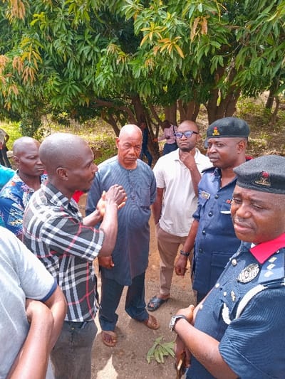 The image shows a group of six men standing in a clearing in a mango orchard in Ungwan Tanko, Nigeria, located at coordinates 10.422 latitude and 7.49 longitude. The men are all dressed casually, and they are interacting with each other. Two of the men are wearing official looking uniforms with a police badge. Two of the men are wearing sunglasses. The men are standing on a dirt path. There is a large mango tree in the background. A leaf is in the foreground, on the dirt path.