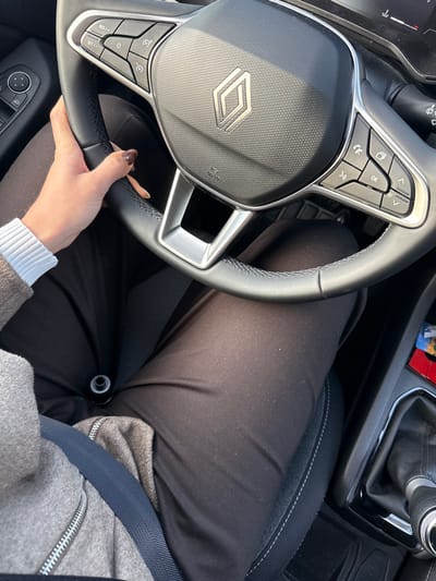 A person is seated in a car. The steering wheel, with a distinctive Renault logo, is held by a hand with painted nails. The person is wearing dark pants, a grey jacket, and a white-cuffed top. A seatbelt is fastened across their body. The car's interior features black seats with white stitching and a black gear shift. The lighting suggests a bright, sunny day.