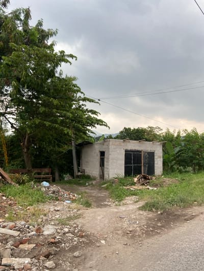 The image shows a small, unfinished concrete building in the middle of a dirt path. The path is located in the city of Kecamatan Margaasih, Indonesia, at coordinates -6.964 latitude and 107.539 longitude. The building has a single door and a window covered with wooden planks. There are several trees and plants in the background, including a large tree on the left side of the image. The ground is covered in dirt, rocks, and debris. The sky is overcast and gray. There are power lines running across the top of the image. There are no people in the image. There is a wooden bench in the image.