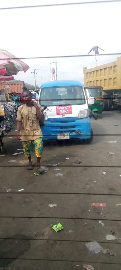 A man in a tan shirt and green and yellow patterned pants is walking towards a blue van parked on a paved street in Igrita, Nigeria (latitude: 4.957, longitude: 7.012). The van has a red sign on the windshield that says "IGWURUTA ENEKA." The van has a license plate that reads "ABG 65XC" and a "FIRST" sticker on the side. The man is looking down at his feet. The street is littered with trash and debris, including a green and white cigarette pack and a red piece of paper. A large yellow dump truck and a green and yellow auto-rickshaw are parked nearby. There are power lines and a solar panel visible in the background. The sky is overcast. The image appears to have been taken through a window with a wire mesh grid.
