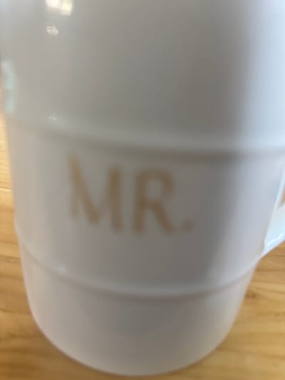 A white mug with the letters "MR." faintly printed on it is shown in the image. The mug is positioned on a light-colored wooden surface. The background is blurred. There are no people or animals present. The location is not discernible from the image.