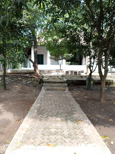 The image shows a paved pathway leading to a set of concrete stairs in front of a white building. The building has a green door and windows. The pathway is surrounded by trees and there is a trash can on the right side of the frame. There are fallen leaves on the ground. This scene is located at the coordinates -6.737 latitude and 108.543 longitude in Cirebon, Indonesia. The scene is likely captured in the afternoon as the sun casts shadows over the pathway. There are no people present in the image. The scene is one of tranquility and solitude, and the white building and trees create a serene atmosphere.