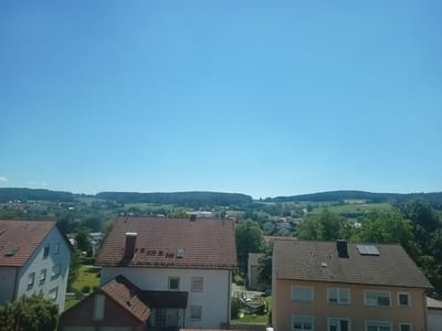 This is an eye-level, wide shot of the city of Oberviechtach, Germany, on a clear, sunny day. The foreground is dominated by the roofs of several houses. On the left, a light-colored, multi-story building with white window frames and a grey tile roof is visible. To its right, a house with a red tile roof and a chimney stands out. In front of this, a smaller section of a building with a dark red tile roof is partially visible. The right side of the image features another building with a brown tile roof, featuring a dark chimney and a solar panel. Beyond the houses, a sprawling green landscape unfolds with rolling hills covered in trees and some scattered buildings. The sky is a clear, vibrant blue with a faint contrail from an aircraft visible in the upper center. There are no people visible in the image, and the scene appears to be a residential area with a backdrop of natural scenery.