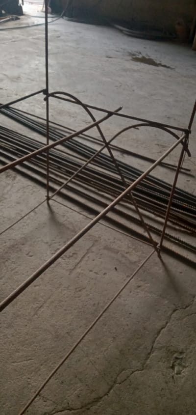 Location: Sumput, Indonesia.
The image shows a set of rusty reinforcing bars (rebar) on a concrete floor. A pre-assembled rebar structure, potentially for concrete reinforcement, stands in the foreground. The structure includes vertical bars and curved horizontal supports. A bundle of straight rebar pieces lies on the floor in the background. There is an area with spilled liquid on the concrete floor. The overall setting suggests a construction site or workshop.