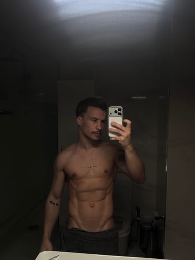 A male individual is depicted in a mirror selfie taken within a bathroom, located in Viseu, Portugal. The man, appearing to be in his twenties or thirties, has dark, side-parted hair, a dark mustache, and a muscular physique with defined abdominal muscles. He is bare-chested, revealing a tattoo of the word "IMPERFECT" (reversed) across his upper sternum and another tattoo (text appearing as "man" reversed) on his upper left arm. He wears a dark grey towel wrapped around his waist. In his right hand, he holds a white smartphone with a light-colored case and a triple-lens camera array, positioned to capture his reflection. The bathroom environment, visible in the reflection, is dimly lit with dark walls, and includes a white toilet and a dark-framed shower enclosure. The edge of a white sink or countertop is visible in the foreground.