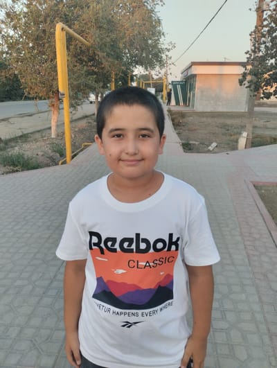 In Yangibazar, Uzbekistan, a young boy stands in a park or pedestrian area, posing for a photo. He is wearing a white Reebok t-shirt with a colorful graphic featuring mountains and the text "Reebok CLASSIC" and "VETUR HAPPENS EVERY WHERE". The boy has dark hair and is looking directly at the camera with a slight smile. He is standing on a pathway made of patterned paving stones. Behind him, there are trees, a yellow pole, and a building with an "ATM 24/7" sign. The environment appears to be outdoors, possibly in the late afternoon or early evening, given the lighting.