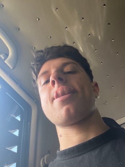 A low-angle, close-up selfie shows a young Caucasian male with dark hair and a light complexion. He is indoors, possibly in a vehicle, as evidenced by the ceiling lining with small, reflective circular studs, a white handle on the left, and a dark tinted window with triangular reflections on the far left. The man is wearing a black t-shirt. His tongue is sticking out, and he is looking slightly upwards. There is a small reddish-brown mole visible on his neck, just below his jawline. The lighting suggests it might be daytime, with some sunlight filtering through the window. The image is sharp and focused on the man's face. There is no information in the image that indicates the location in the city of Pechinska, Bulgaria.