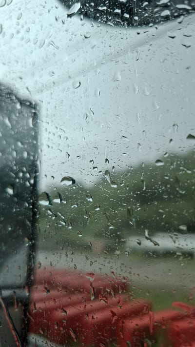 The image shows a view through a wet window, with raindrops distorting the scene outside. The foreground is dominated by the raindrops on the glass, which are of various sizes and shapes, some creating streaks as they have run down. In the background, the blurry shapes of objects and a landscape can be seen. On the left side of the image, a dark, vertical structure is visible, likely part of a vehicle or building. Below this, a bright red object or structure is partially visible, appearing textured. Further in the background, a green, blurred area suggests trees or foliage, and beyond that, a lighter, overcast sky. The overall impression is one of a rainy day, with limited visibility due to the weather and the water on the window. The location in Budukan, Russia is not discernible from the image itself.