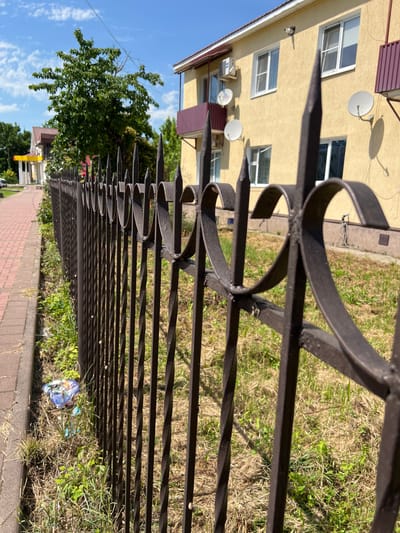 The image shows a wrought iron fence in front of a yellow building with a reddish-brown roof. The fence has decorative swirls and pointed spikes. Beyond the fence, there is a paved walkway with red bricks, and beyond that, a green tree and another building. The ground next to the fence is covered in dry grass and some green weeds, with a small pile of colorful trash visible in the lower left corner. The sky is blue with scattered white clouds. The building has several windows, air conditioning units, and satellite dishes. The overall scene suggests an outdoor environment in a residential or commercial area.
