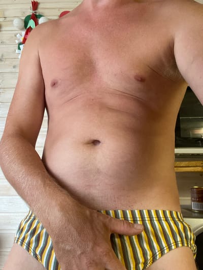The image shows a close-up, waist-up view of a shirtless person wearing striped briefs. The person's left arm is resting on their lap, obscuring part of their lower abdomen. The background appears to be a wooden wall with some Christmas decorations on the left. The lighting suggests an indoor setting. There is no visible information in the image that indicates a specific location in Narynka, Russia.