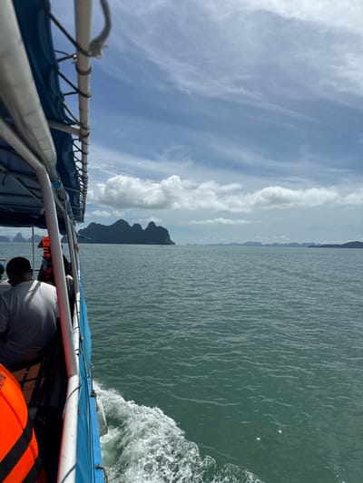 An eye-level shot captures a boat sailing on the teal-colored ocean, heading towards a cluster of rocky islands in the distance. The boat, a turquoise and white vessel with a blue canopy, is in the foreground, with a wooden deck visible. Several passengers are seated on the deck, some wearing life jackets. One person in a white shirt is looking out at the sea, while another person in an orange life jacket stands. The water is calm, with a visible wake trailing from the boat. The sky is partly cloudy, with scattered white clouds against a light blue backdrop. The islands in the distance are dark and mountainous, with a hazy horizon. The scene appears to be from the viewpoint of someone on the boat, looking forward and slightly to the right. The location is identified as Khlong Khian, Thailand.