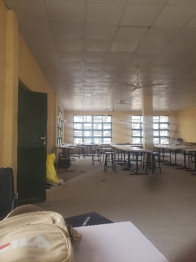 This image depicts the interior of a large, empty classroom or laboratory facility located in Kaninkon, Nigeria. The room features light yellow-beige walls and a greyish concrete or tiled floor. A suspended ceiling with square panels shows signs of wear and discoloration. Natural light illuminates the space through multiple large, barred windows along the back wall. Several dark-framed tables with accompanying dark stools are arranged in rows across the room. Three ceiling fans are visible overhead, and air conditioning units are mounted below some windows. A structural pillar stands towards the center-right of the room. A dark green door, slightly ajar, is positioned on the left side of the frame, with a crumpled yellow object resting on the floor nearby. In the immediate foreground, a light-colored Fila-branded backpack rests on a white surface, partially obscuring a dark notebook or folder with "TETFund" visible on its spine, alongside a blank white sheet of paper. No individuals are present in the scene.