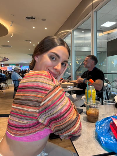 A brightly lit, slightly high-angle interior shot shows a woman with brown hair and fair skin looking towards the camera. She rests her cheek on her hand. She wears a colorful striped top with a pink lace hem. Behind her, a man with short hair in a black shirt is seated at a table, seemingly looking at something in front of him. On the table is a Starbucks iced coffee and what appears to be an iced tea drink, along with a blue plastic bag and a small takeout container. In the background, a food court area with several businesses is visible, including a "CINNABON" sign. The location is identified as Spata-Loutsa Municipal Unit, Greece.