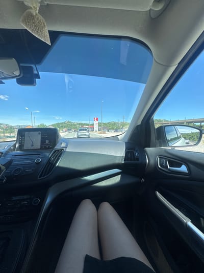 The image shows the interior of a car driving on a sunny day, likely on a highway or main road. The view is from the passenger seat, looking forward and to the right. The dashboard is visible with a large touchscreen display showing a navigation map. To the right, the side mirror reflects a road and trees. In the foreground, a person's legs are visible, resting on the passenger seat, suggesting they are a passenger in the car. The road ahead is lined with trees and some buildings, and there are other cars on the road. In the distance, hills and mountains are visible under a clear blue sky. A gas station with red and white branding is also visible on the right side of the road. Based on the presence of the EKO gas station, this location is likely near Očinići, Montenegro. A small, clear bag filled with small white beads, possibly an air freshener, hangs from the car's ceiling.
