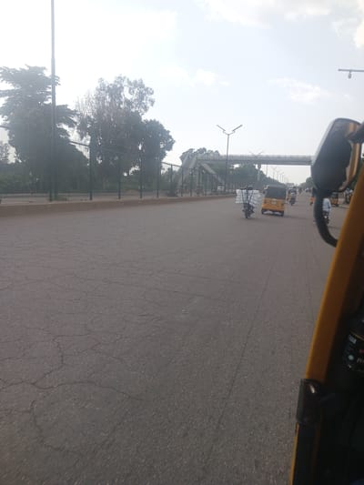 The image depicts a street scene in Unguwan Dosa, Nigeria, captured from the perspective of a moving vehicle. The road is paved with asphalt and shows signs of cracking. On the left side of the road, there is a metal fence and lush green trees, with a tall pole extending upwards. A pedestrian bridge is visible in the distance, spanning across the road. Several vehicles are present, including auto-rickshaws (Keke Napep) and motorcycles. One auto-rickshaw in the foreground is yellow, and another further down the road is also yellow. A motorcycle in front of the yellow auto-rickshaw is carrying a load of white rectangular objects. Another auto-rickshaw further ahead is yellow. In the far distance, more vehicles and structures can be seen. The sky is overcast with clouds.