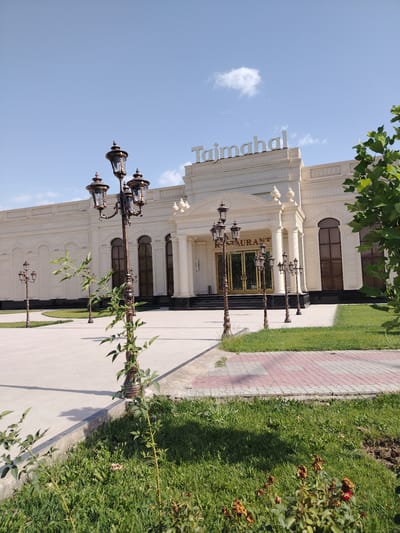 The image shows the Taj Mahal Restaurant in Shahrisabz City, Uzbekistan, under a clear blue sky with a few wispy clouds. The building is a large, cream-colored structure with ornate columns and arched windows. A sign above the entrance reads "Taj Mahal Restaurant." In front of the building is a wide, paved walkway lined with decorative lampposts. Green grass and some small plants are visible in the foreground and along the sides of the walkway. The scene appears to be calm and the area is well-maintained.
