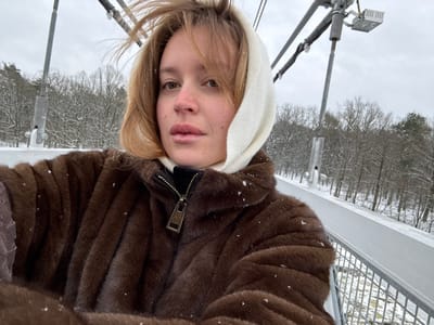 The scene depicts an outdoor winter environment on a suspension bridge in Мастоўскі сельскі Савет, Belarus. A female individual, appearing to be an adult, occupies the foreground in a close-up, self-portrait-style photograph. She has light-colored hair, partially covered by a cream-colored headscarf or hood. Her face is visible, with a neutral expression directed towards the camera. She wears a brown faux fur coat with a black collar featuring a brass-toned zipper pull. Small, distinct snowflakes are present on her clothing and hair, indicating active or recent snowfall.

The background reveals elements of a metallic suspension bridge, including grey support cables, vertical struts, and a snow-covered white railing. A cylindrical metallic post and a square light fixture are attached to the bridge infrastructure in the upper midground. Beyond the bridge, a dense, leafless forest covered in snow extends into the distance. The ground below the bridge and throughout the forest is uniformly covered in snow. The sky is overcast and grey.