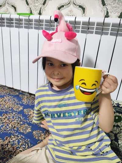 A young child, likely a girl, is pictured sitting on a patterned blue carpet in front of a white radiator. She wears a pink baseball cap adorned with a plush flamingo toy. The cap has the word "Sweet" written on it in pink lettering. She is also wearing a horizontally striped t-shirt with yellow and light purple stripes, and the text "HARVESTER" and "WE LACK TO DISCOVERY" is visible on the shirt. In her right hand, she holds a bright yellow mug decorated with a laughing emoji face. The child is looking towards the camera with a slight smile. The background features a decorative wall with a repeating patterned wallpaper and the white radiator. The lighting suggests an indoor setting. The location context is Навруз, Uzbekistan.