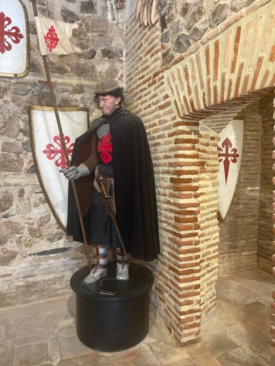 This is a photograph of a life-size mannequin dressed as a medieval knight, standing inside a stone and brick structure. The knight is holding a wooden pole with a tattered white flag adorned with a red emblem. He wears a dark cape, a chainmail shirt, a helmet, gloves, and armored boots. A sword is sheathed at his side. To the left and right of the knight are large shields, each featuring a red emblem on a white background. The background consists of rough stone walls and an arched brick entryway. The floor is paved with stone tiles. Based on the emblem on the flag and shields, which appears to be the Cross of Saint James, this scene is likely located at the Alcázar of Toledo, a historic fortress and palace in the city of Toledo, Spain.