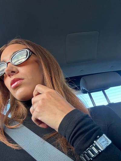 This image depicts a female subject seated inside a vehicle in Villers-Agron-Aiguizy, France, during daylight hours. The subject, visible from the upper torso, wears a black long-sleeved garment and a gray fabric seatbelt diagonally across her chest. Her reddish-brown hair extends over her shoulders. She is equipped with oval, tortoiseshell-patterned sunglasses that reflect a sky with scattered clouds. Her lips are glossed. A silver-toned ring is present on her left ring finger. On her left wrist, a rectangular silver-toned wristwatch with a metal link band, featuring a light blue face and Roman numerals (consistent with a Cartier Tank Française model), is worn. Her left hand is elevated, with fingers resting against her chin. The vehicle's dark gray headliner is visible above, and a portion of a gray fabric headrest is seen behind the subject. A clear, blue sky is visible through the vehicle's rear window.