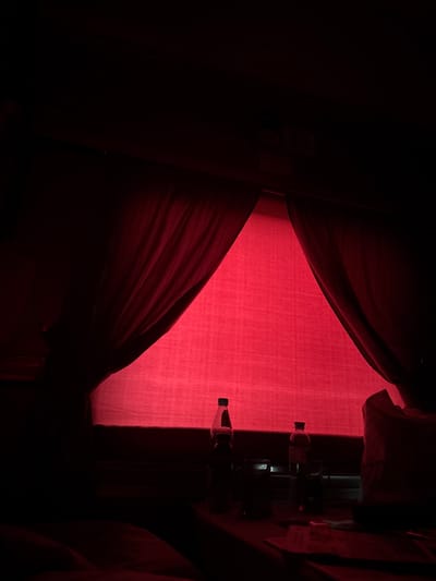 The image depicts a dimly lit scene with a prominent red-hued fabric, possibly a curtain or drape, dominating the frame. The fabric is illuminated from behind, creating a vibrant red glow that casts shadows and highlights the texture and folds. On the right side of the image, a vertical structure, possibly a window frame or a shelf, is visible, holding several objects. These objects appear to be bottles, with their shapes and caps discernible. They are silhouetted against the red light, making their details indistinct. The overall impression is one of mystery and subdued ambiance, potentially set in an indoor environment. The time of day and weather conditions are not discernible. There is no visible text in the image.