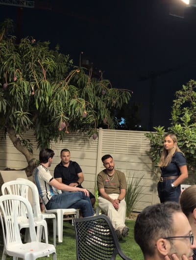 The image depicts a nighttime outdoor gathering at a private residence in Lod, Israel. Four individuals are present in the foreground: three men and one woman. One man, wearing a white and blue striped shirt and blue jeans, is seated. To his right, another man in a dark polo shirt and dark shorts is also seated. A third man, in a green t-shirt and light-colored pants, is seated with his legs crossed. A woman in a denim jumpsuit is standing to the right of the seated men. She is looking towards the right side of the frame. The group is positioned in a backyard setting with artificial turf and a white slatted fence in the background. A tree with visible fruit, possibly mangoes, is prominent on the left side of the image. The overall atmosphere suggests a casual social event.
