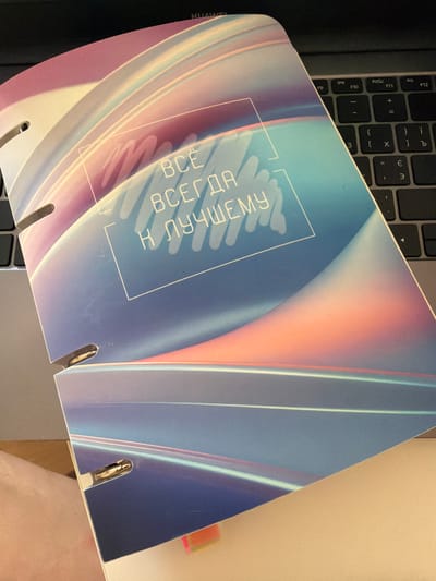 The image shows a close-up, overhead view of a notebook with a holographic, iridescent cover lying on a laptop keyboard. The notebook is angled, with the top left corner in focus and the bottom right corner slightly out of focus. The cover of the notebook features abstract swirling lines in shades of pink, purple, blue, and teal, with a white rectangular outline containing white text in Cyrillic that reads "ВСЁ ВСЕГДА К ЛУЧШЕМУ" (Everything always for the best). The notebook has three metal binder rings visible on its left edge, with the top two peeking out from behind the cover and the bottom one clearly visible. The laptop keyboard is dark gray and partially visible in the background. The overall lighting suggests an indoor setting, likely an office or home environment. There is no indication of the city of Кирпичного завода № 1, Russia, in the image.