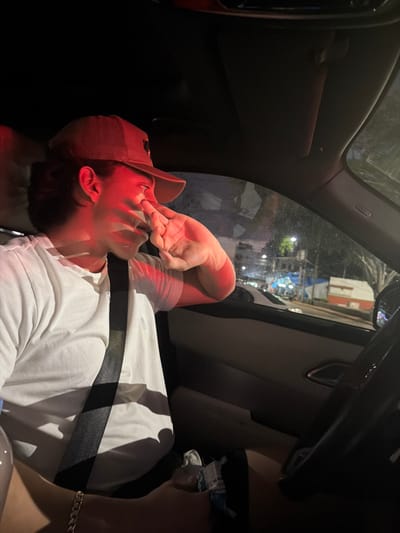 Inside a car, a person wearing a red baseball cap and a white shirt is reclined in the passenger seat with a seatbelt on. Their hand is near their face, specifically their nose. The interior is dark, but through the window, one can see an urban street scene at night. The view shows buildings and streetlights, giving a glimpse of the outside environment.