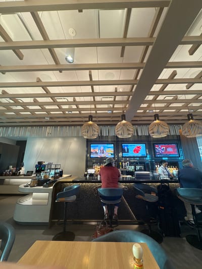 The image depicts the interior of a modern bar or lounge establishment, highly characteristic of an airport lounge. The specific location within the city of Arlington, United States, is not discernible from the visual evidence; however, the decor is consistent with facilities found in major transportation hubs like Dallas/Fort Worth International Airport, which serves the Arlington area.
The scene features a prominent bar counter with a dark, reflective surface and a textured, dark-paneled facade. Three individuals are visible at the bar. In the center, an individual with their back to the camera wears a red shirt and a light-colored wide-brimmed hat, seated on a blue upholstered bar stool. A brown leather bag rests on the floor beneath their stool. To their right, a female bar employee in a white collared shirt and dark apron stands behind the bar, appearing to prepare drinks amidst numerous liquor bottles and bar equipment. To her right, a second patron in a dark jacket is seated at a bar stool, partially visible.
Above the bar, four large, circular pendant lights with slatted wooden shades illuminate the area. Behind the bar, a metal frame supports three large flat-screen televisions. The left screen displays a cooking show or lifestyle program. The middle screen shows what appears to be a news or information graphic with a red outline. The right screen displays a sports event, specifically college basketball ("NCAAB" visible in the corner graphic).
To the left of the bar, a contemporary, light-colored service counter area features multiple coffee machines, beverage dispensers, and clear containers holding snacks or pastries. The walls are smooth, light-toned, and feature subtle paneling. The ceiling is a distinctive architectural element, comprising a grid of light-colored wooden beams with white panels and integrated recessed lighting fixtures and speaker grilles. In the immediate foreground, a light-colored wooden tabletop and the back of a curved, dark blue chair are partially visible, with a small decorative bottle on the table. The floor is not clearly discernible.