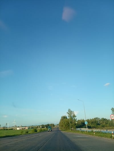 The image depicts a road scene in the vicinity of Вяйнянскі сельскі Савет, Belarus. The perspective is from a vehicle moving along a paved road. On the left side of the road, there is a grassy field with several wind turbines visible in the distance, along with some low-rise buildings. A green truck is visible on the road ahead, traveling in the same direction as the viewer. On the right side of the road, there is a body of water bordered by a metal guardrail, and a dense line of trees. A street light and a blue road sign indicating a directional change are also present on the right side of the road. A circular speed limit sign with the number "60" is also visible on the right. The sky is clear blue with a few faint clouds. The overall impression is of a rural or semi-rural landscape during daylight hours.