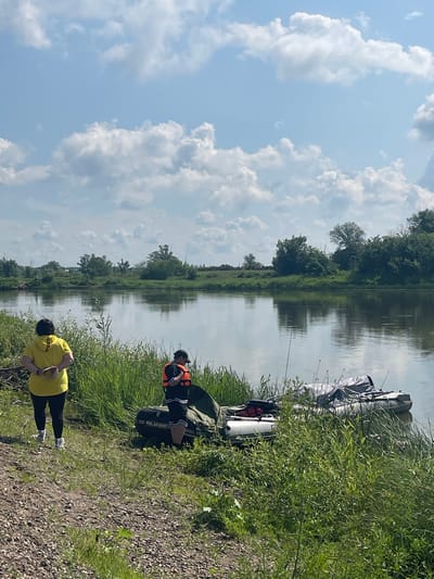 The image depicts a serene river scene with two individuals standing on the grassy bank. In the foreground, a person wearing a bright yellow hoodie and black leggings stands with their hands clasped behind their back, facing away from the camera. To their right, another individual in a black t-shirt with white stripes and an orange life vest is positioned near an inflatable boat. The boat, a "Polar Bird," is partially covered with a grey tarp and rests by the water's edge. A second, smaller inflatable boat is also visible. The river is calm, reflecting the lush green trees and a partly cloudy sky. The background shows a dense line of trees and vegetation along the opposite bank, with more trees and a field visible in the distance. The overall impression is of a tranquil, outdoor setting, likely a recreational area near Nezhinka, Russia.
