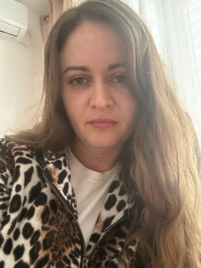 A woman with long brown hair is facing the camera. She is wearing a white t-shirt and a brown and white animal print jacket with a zipper. The background appears to be indoors. It is impossible to confirm the location as Karmiel, Israel without further information.