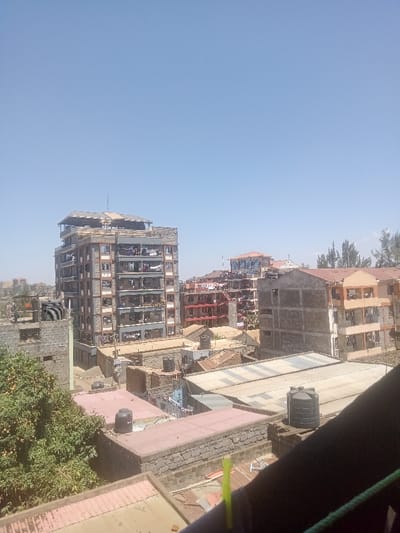 This image depicts a dense urban landscape in Gatongora ward, Kenya, under a clear, bright blue sky. The foreground features several rooftops, some flat and concrete-like with visible black cylindrical water tanks, others with corrugated metal sheeting. In the lower-right corner, a dark, diagonal obstruction suggests a railing or architectural element from the vantage point of the photographer, with a distinct yellow-green cylindrical object (possibly a straw or cable) and a green rope visible.
The midground is dominated by a prominent multi-story building on the left, constructed with visible concrete blocks and having numerous balconies on its lighter-colored upper section and darker lower section. Clotheslines with garments are visible on many balconies. The uppermost part of this building shows ongoing construction or an unfinished rooftop structure with metal scaffolding. To the right of this central building, the cityscape continues with more multi-story residential or commercial buildings of varying architectural styles and heights. Many of these structures also display clothes drying on balconies or rooftops.
In the background, the urban environment extends further with more buildings. Sparse trees with green foliage are present, particularly in the lower-left portion of the image. No individuals are distinctly identifiable. The overall impression is that of a bustling, residential or mixed-use urban area during daytime.