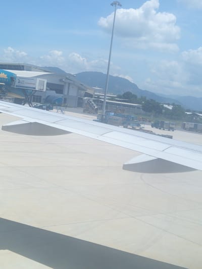An aerial view of an airport tarmac in Bắc Cam Ranh, Vietnam shows an airplane parked at a gate, with a jet bridge extended to its door. The plane is blue and yellow with a flower emblem on its side. In the background, there is a terminal building, mountains, and a bright blue sky with scattered clouds. Several service vehicles, including a luggage tug and a fuel truck, are visible on the tarmac. The wing of another aircraft dominates the foreground, casting a shadow on the tarmac.