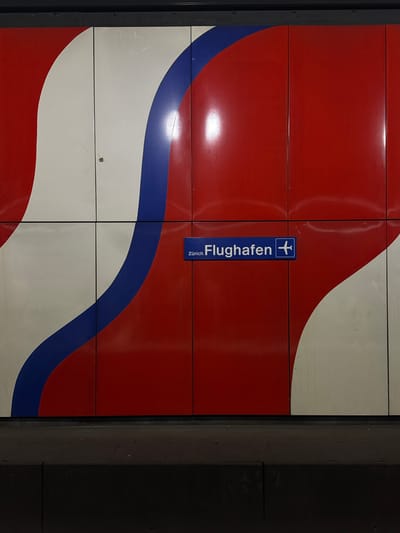 The image displays a section of a tiled wall and platform edge, likely within an underground train or airport terminal in Kloten, Switzerland, specifically at Zürich Airport. The wall is composed of rectangular, glossy panels featuring a distinct, curvilinear design in red, white, and blue. A wavy blue stripe separates larger sections of red and white, which also have undulating boundaries. A prominent rectangular blue sign with white lettering, "Zürich Flughafen," and a white airplane pictogram is mounted on a red panel section in the central-right area of the wall. The glossy surfaces of the panels exhibit reflections of overhead lighting. A small impact mark, characterized by a brown discolored spot and radiating cracks, is visible on a white panel in the upper-left quadrant. Below the wall, a dark, elevated platform surface extends across the lower portion of the frame. No people or active events are present in the scene.